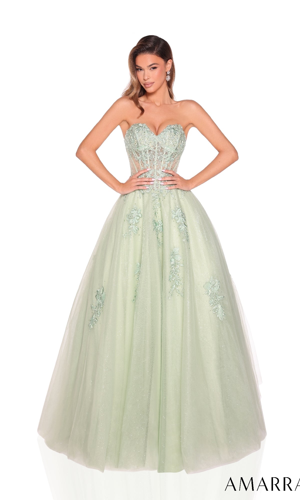 A woman wears the Amarra 89011 Lace-Up Strapless Prom Ball Gown in light green, featuring floral embellishments and a fitted, beaded sweetheart bodice, set against a white background.